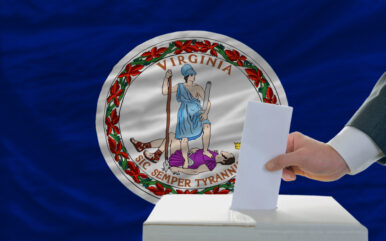 man putting ballot in a box during elections in front of flag american state of virginia