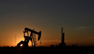 A pump jack operates in front of a drilling rig at sunset in an oil field in Midland